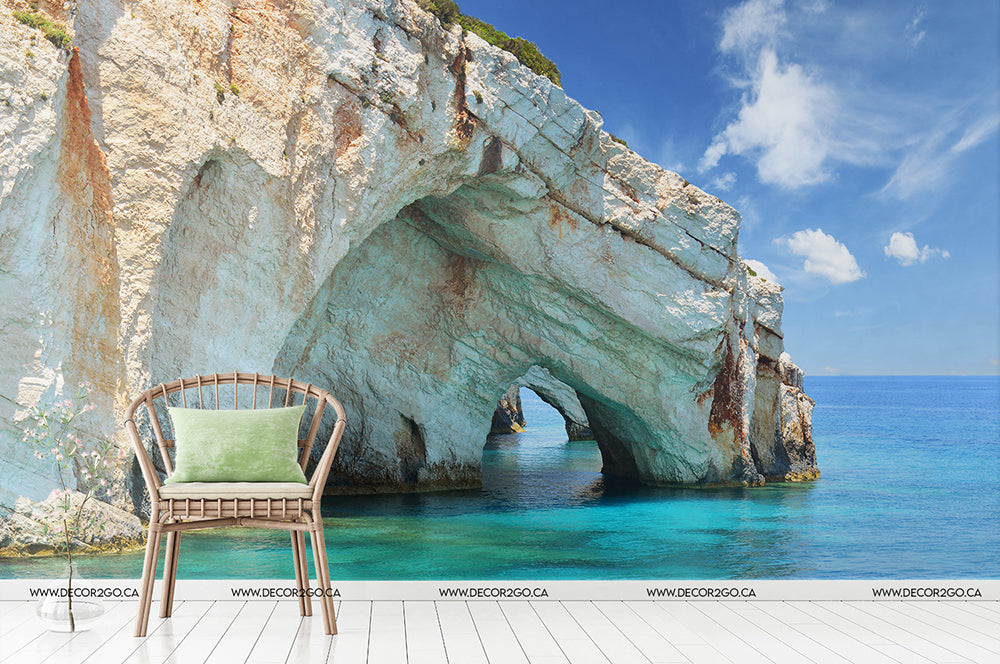A wicker chair with a green cushion rests on a white wooden floor, set against the Decor2Go Wallpaper Mural's Aquatic Cavern design, which showcases a rocky coastal archway with blue ocean water and a clear sky.