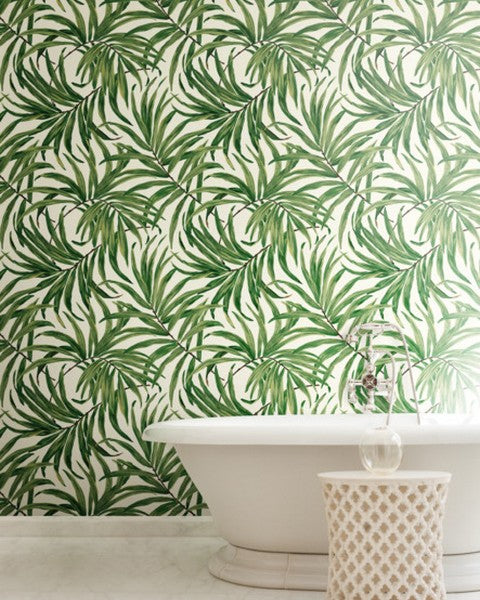 A contemporary white freestanding bathtub against a wall with vibrant York Wallcoverings Bali Leaves Wallpaper (60 SqFt). A small white waste basket and a clear bottle are beside the tub.