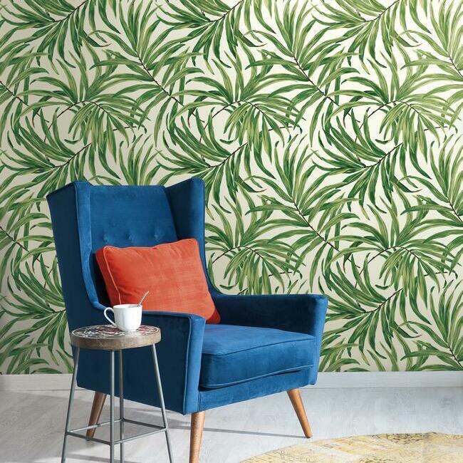 A cozy room featuring a blue armchair with an orange pillow, beside a small round table holding a white mug. The wall is adorned with the Green Bali Leaves Wallpaper (60 SqFt) from York Wallcoverings, infusing the space with a nature-inspired design through its green and beige tones.