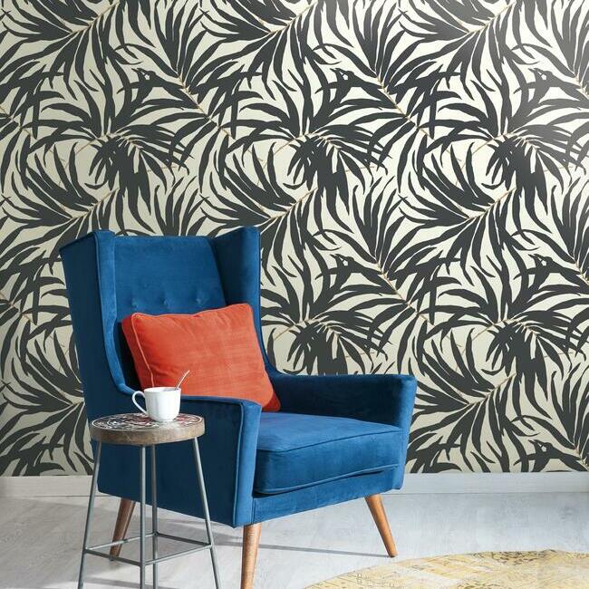 A vibrant blue armchair with an orange cushion next to a small table with a cup, set against the bold York Wallcoverings Bali Leaves Wallpaper (60 SqFt).