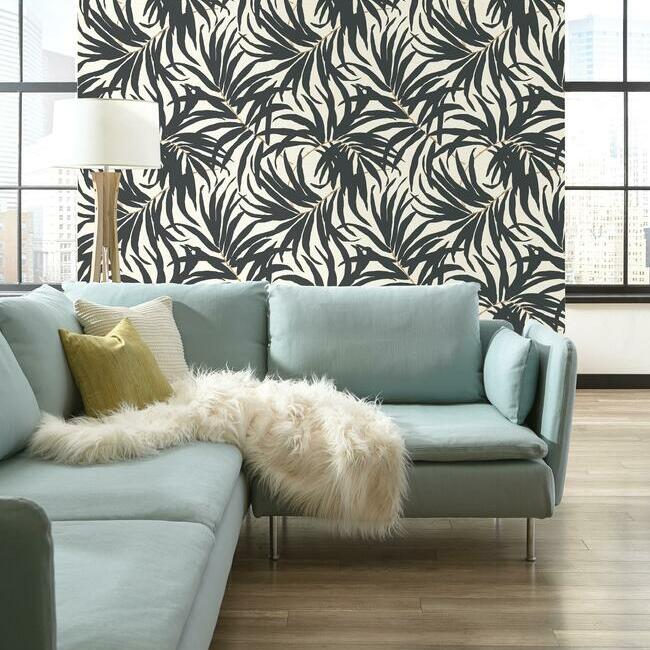 A stylish living room with a light blue sectional sofa adorned with a white fur blanket and yellow pillow, set against a bold black and white York Wallcoverings Bali Leaves Wallpaper (60 SqFt).