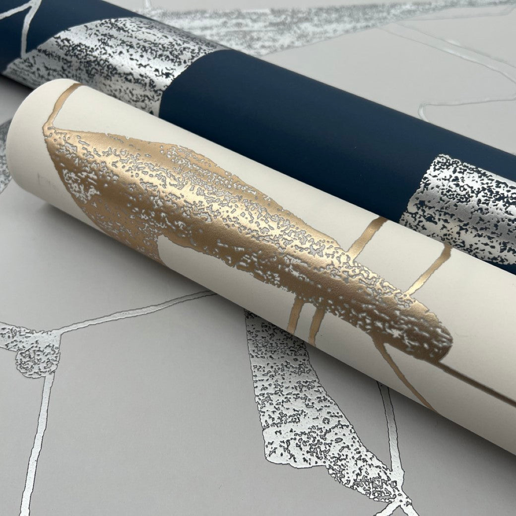 Displayed are two rolls of wallpaper: one dark blue with silver accents and another, the Cream & Gold Abstract Geo Wallpaper by York Wallcoverings, featuring beige with gold and white patterns. Part of the Modern Metals Collection, they offer a modern design with geometric metallic flair.