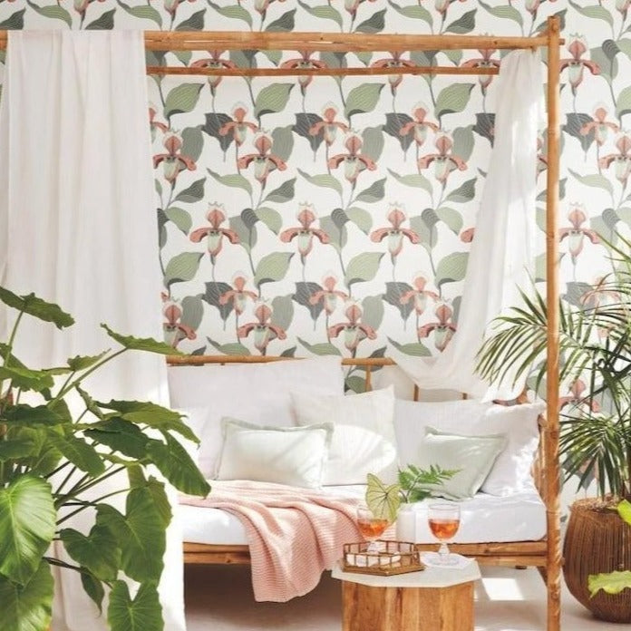 A cozy bedroom with York Wallcoverings' Lady Slipper wallpaper, featuring a wooden canopy bed draped with white sheer curtains, surrounded by lush green plants and a small table with drinks.