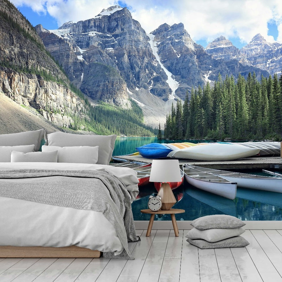 A bedroom with a bed and side table next to the Decor2Go Wallpaper Mural, seamlessly blending into a scenic landscape featuring a crystal-clear lake, kayaks, and towering mountains in the background.