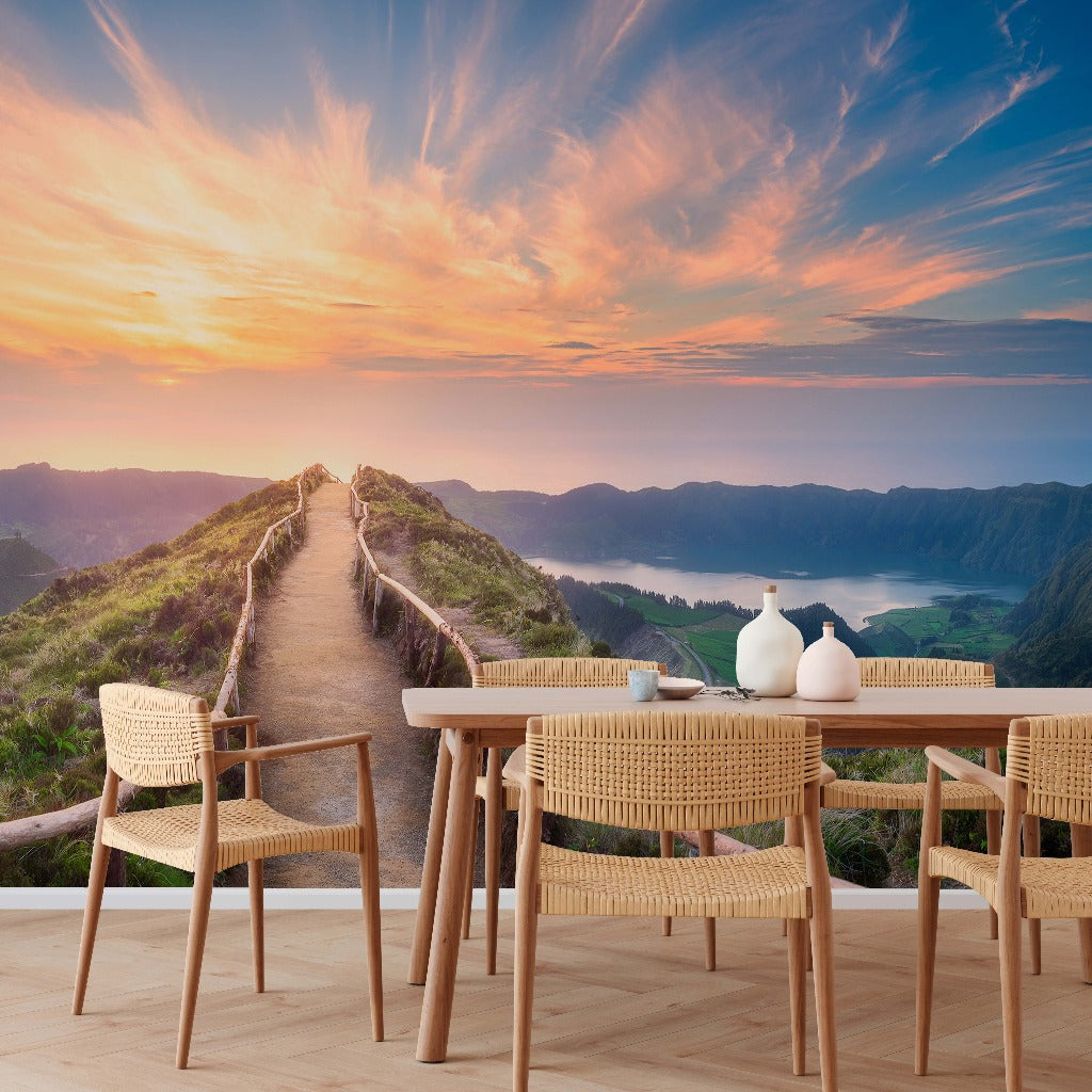 Outdoor dining setup with rustic wood furniture and wicker chairs overlooking a Mountaintop Trail Wallpaper Mural from Decor2Go Wallpaper Mural leading through lush green hills under a vibrant sunset sky.