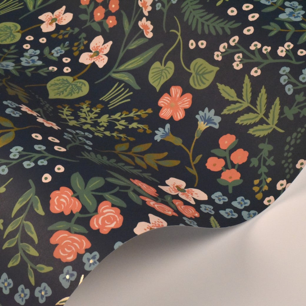 A sheet of paper with a dark background showcases York Wallcoverings’ Navy Wildwood Wallpaper (60 SqFt), featuring a vibrant floral pattern in pink, orange, blue, and white flowers with green leaves, inspired by Rifle Paper Co’s design.
