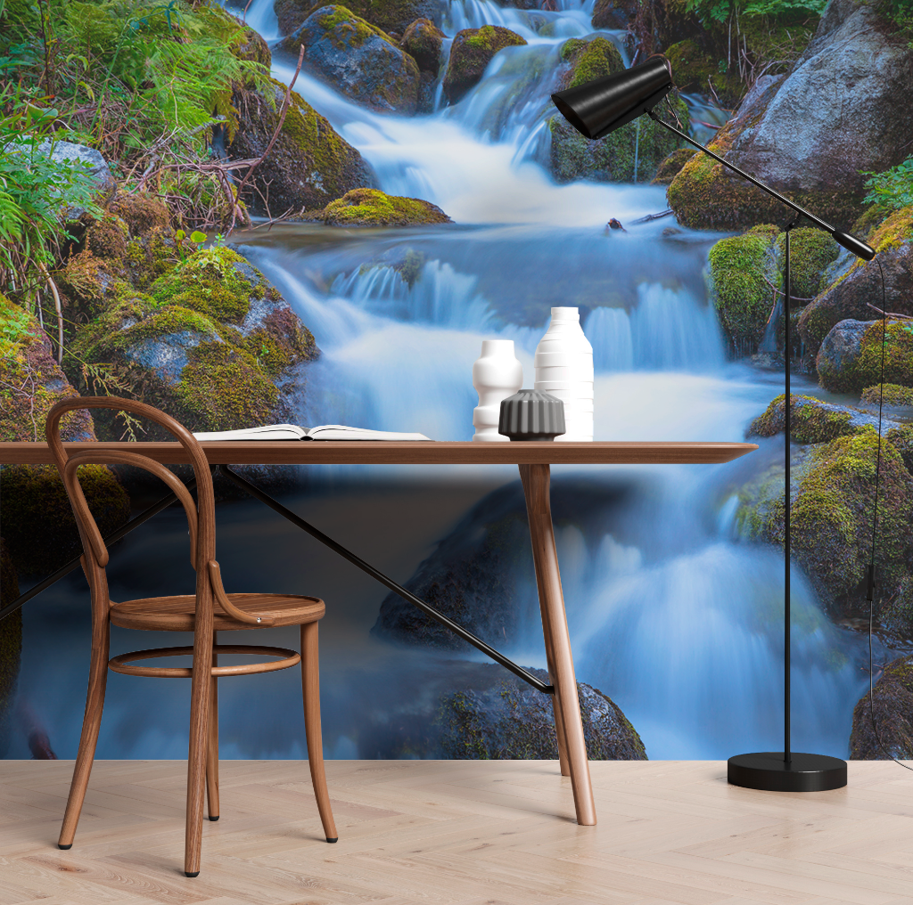 A serene home office setup with a wooden chair and desk against a large, vivid nature mural of a Decor2Go Wallpaper Mural Peaceful Waterfall Wallpaper Mural surrounded by mossy rocks, complemented by a black desk lamp and white decorative vase.