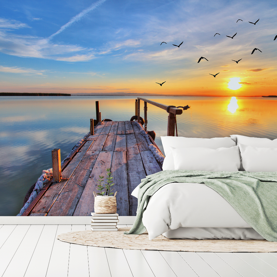 A serene bedroom seamlessly blending into a tranquil lakeside retreat, featuring a bed with white bedding on a wooden deck leading to a small pier, with the "Sunset Birds Flying Wallpaper Mural" by Decor2Go Wallpaper Mural in the background.