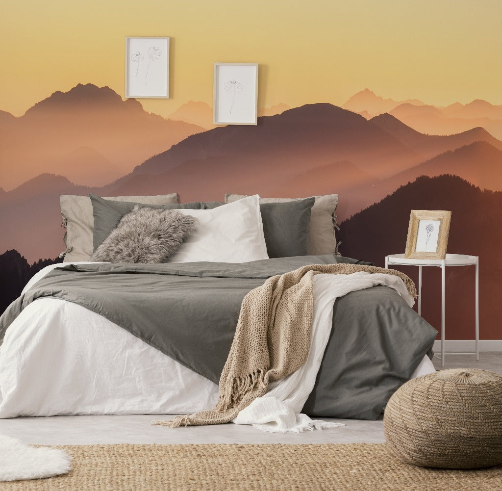A cozy bedroom setup featuring a neatly made bed with white and olive green bedding, a knit throw, and fluffy pillows. The backdrop shows a Decor2Go Wallpaper Mural of Sunset on the Mountains during sunset. Decorative elements include framed art and a floor cushion.
