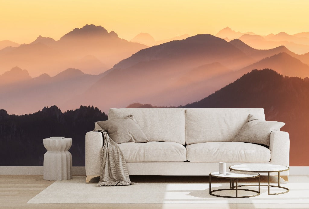 A modern living room setup with a beige sofa, small tables, and a rug, overlooking a tranquil Sunset on the Mountains Wallpaper Mural bathed in the warm glow of a sunset from Decor2Go Wallpaper Mural.