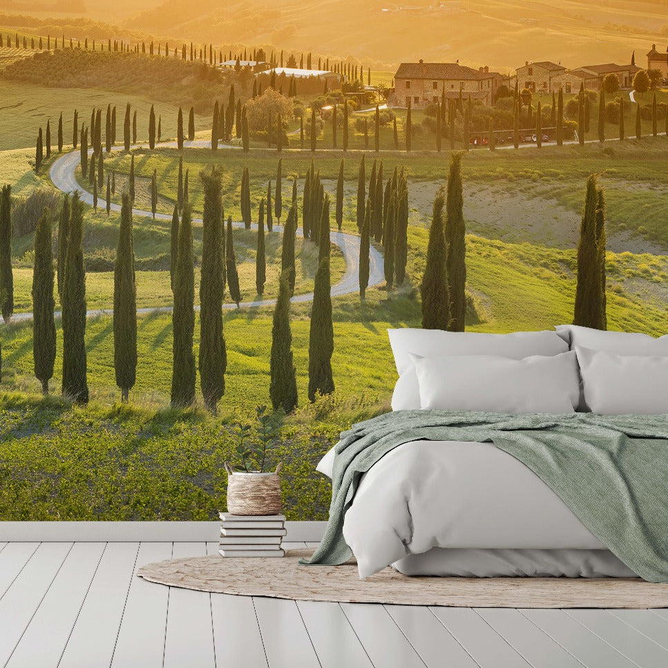 A modern bedroom with a large bed facing a wall-sized, realistic Decor2Go Toscana Trail Wallpaper Mural depicting vibrant green hills, a winding road, and rows of slender cypress trees under an orange sunset.