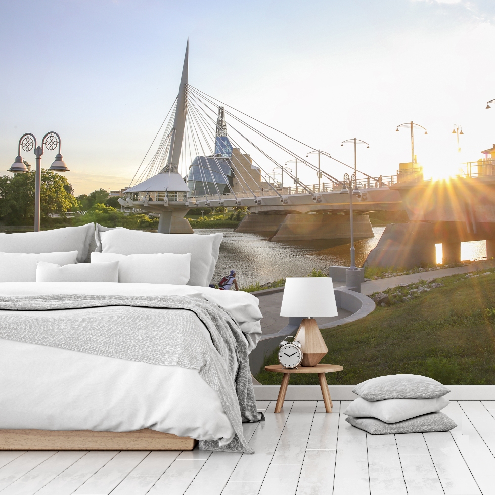 A surreal image of a cozy bedroom setup, including a bed and bedside table, placed outdoors on a wooden deck with a scenic view of modern architecture Winnipeg over a river at sunset featuring the Decor2Go Wallpaper Mural Urban Bridge Wallpaper Mural.
