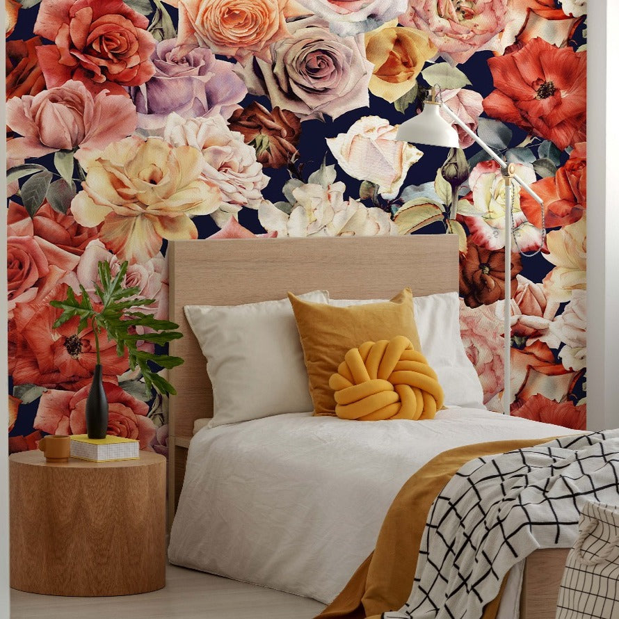 A cozy bedroom featuring a large bed with white bedding and yellow accents in front of a vibrant Decor2Go Wallpaper Mural with various colorful roses. A wooden side table with a plant and lamp.