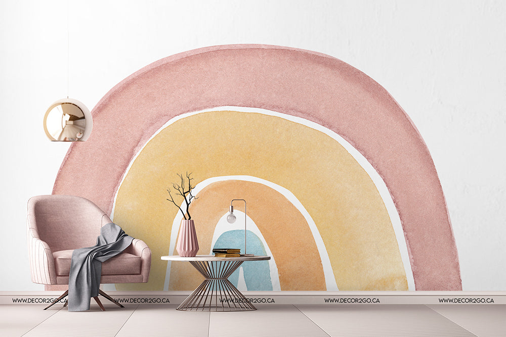 A stylish children's room corner with a Watercolor rainbow Wallpaper Mural arch by Decor2Go Wallpaper Mural, featuring a plush pink armchair, a gray throw blanket, a round side table with a vase and books, and a modern hanging