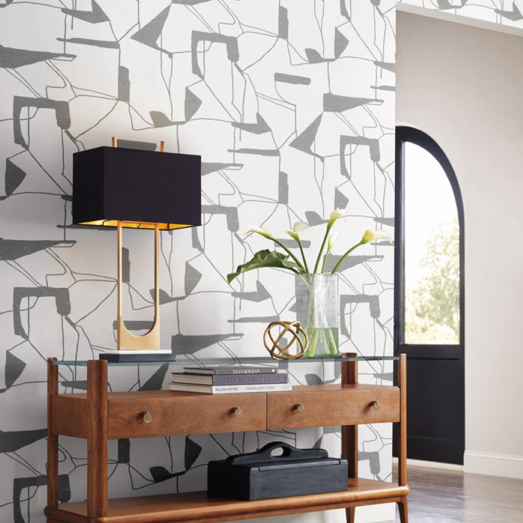A wooden console table with drawers holds a decorative orb, a glass vase filled with green leaves, and a small box. A modern sconce with a black shade is mounted on York Wallcoverings' White & Silver Abstract Geo Wallpaper. An arched window in the background enhances the contemporary design.