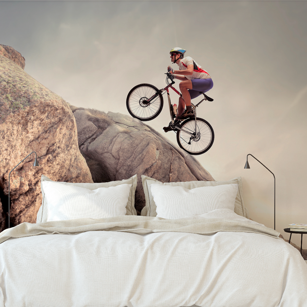 Woman wearing a helmet and sunglasses rides a mountain bike over a bed with white bedding, set against a surreal backdrop of large rocks using the Decor2Go Wallpaper Mural.
