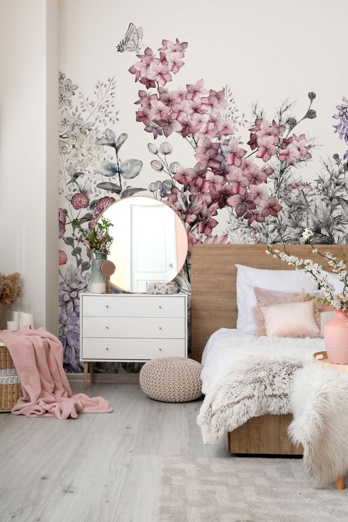 A cozy bedroom with a Wild Flower Wallpaper Mural feature wall, a wooden bed covered with soft white and furry textiles, a small white dresser with a round mirror, and decorative items like a vase and potted plants.