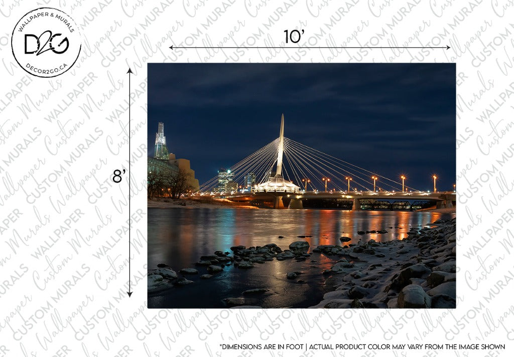 An illuminated bridge with white cables spans over a river at night, reflecting on the water with city buildings in the background, and a rocky riverbank in the foreground. This Winnipeg in Blues Wallpaper Mural creates a romantic atmosphere.