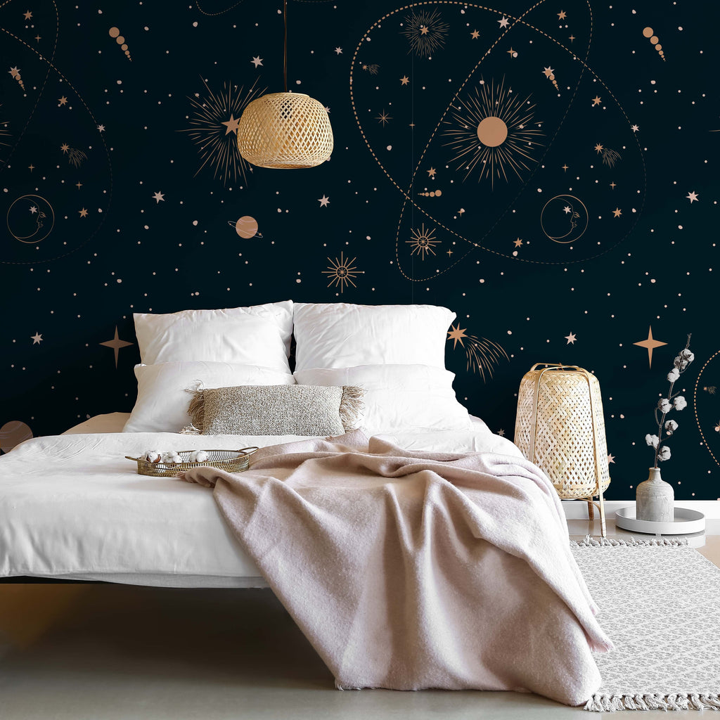 A cozy bedroom with a white bed and beige blanket, against a dark blue Decor2Go Wallpaper Mural featuring planets and constellations. A woven nightstand and lamp complement the scene.