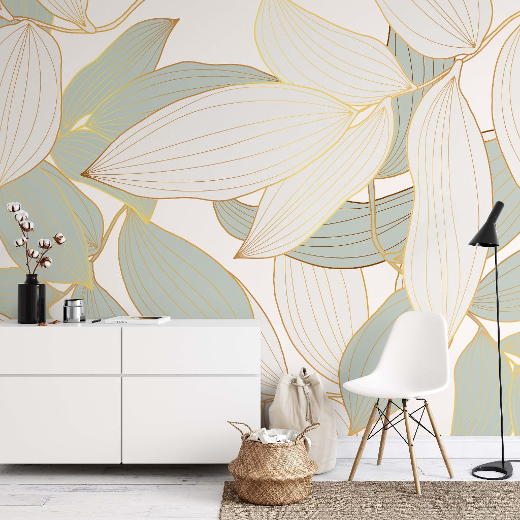 A modern room with a large Decor2Go Wallpaper Mural featuring oversized blue and golden leaves. A white chair, a side cabinet, and decorative elements such as a small plant and a basket are present.