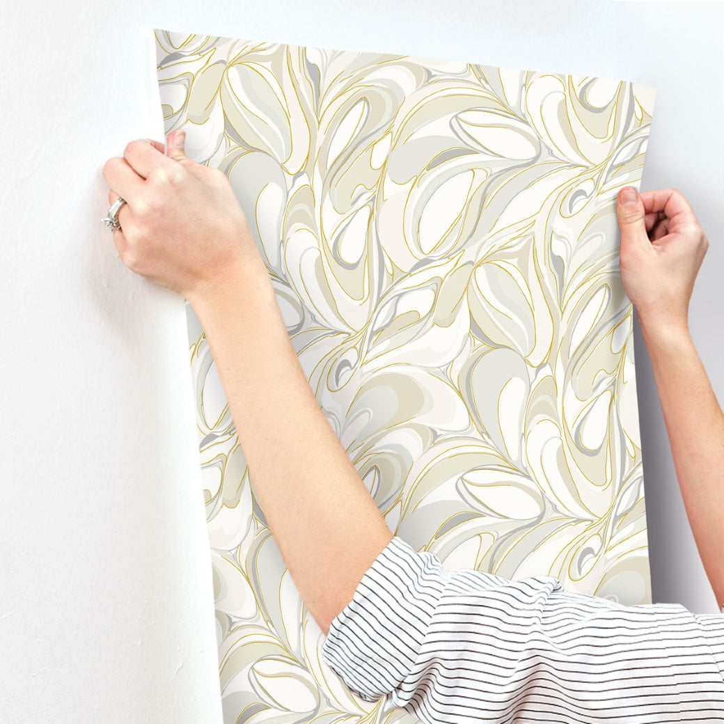 A person in a striped shirt smooths York Wallcoverings' Achate Beige & Grey Wallpaper (60 SqFt), featuring swirling leaf designs and subtle metallic gold veining, onto a white wall.