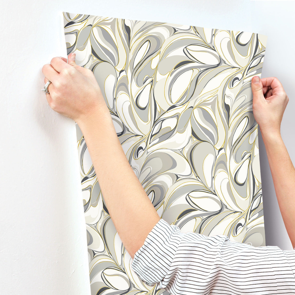 A person with rolled-up sleeves hangs York Wallcoverings' Achate Black & Grey Wallpaper (60 SqFt), featuring a swirling marbled design in shades of black, grey, and white, against a white wall.