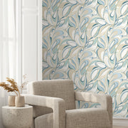 A cozy beige armchair and a small round table with a vase of dried plants sit before a wall adorned with York Wallcoverings' Achate Blue Wallpaper (60 SqFt) featuring blue, green, and cream tones.