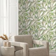 A modern living space features a light gray armchair, a round side table with dried flowers, and York Wallcoverings' Achate Grass Wallpaper in bold green, beige, and metallic gold. Natural light enhances the vibrant leafy design.