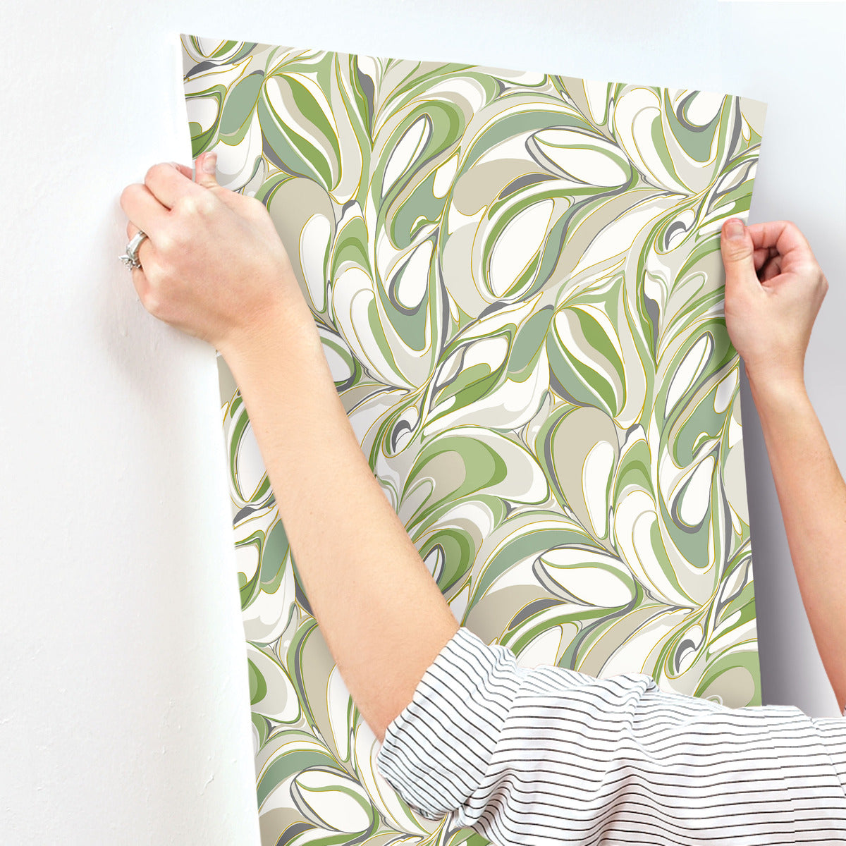 A person in a striped shirt applies York Wallcoverings' Achate Grass Wallpaper, featuring a green and cream marbled leaf pattern, onto a white wall.