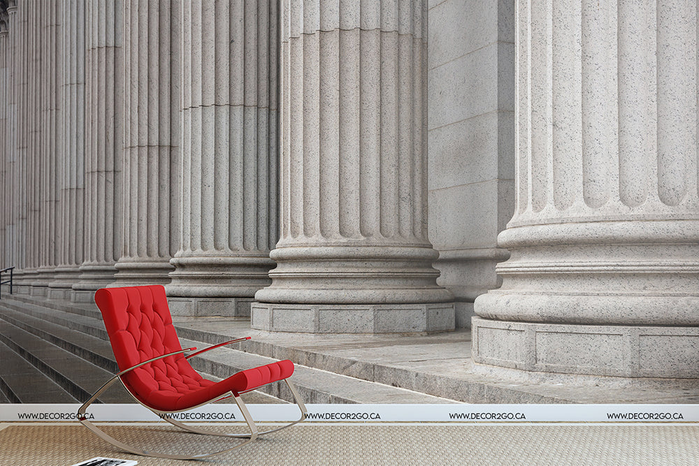 A modern red lounge chair positioned in front of a grand building with imposing Greek architecture, accentuating a striking contrast between the Ancient Pillars Wallpaper Mural by Decor2Go Wallpaper Mural and traditional architecture.