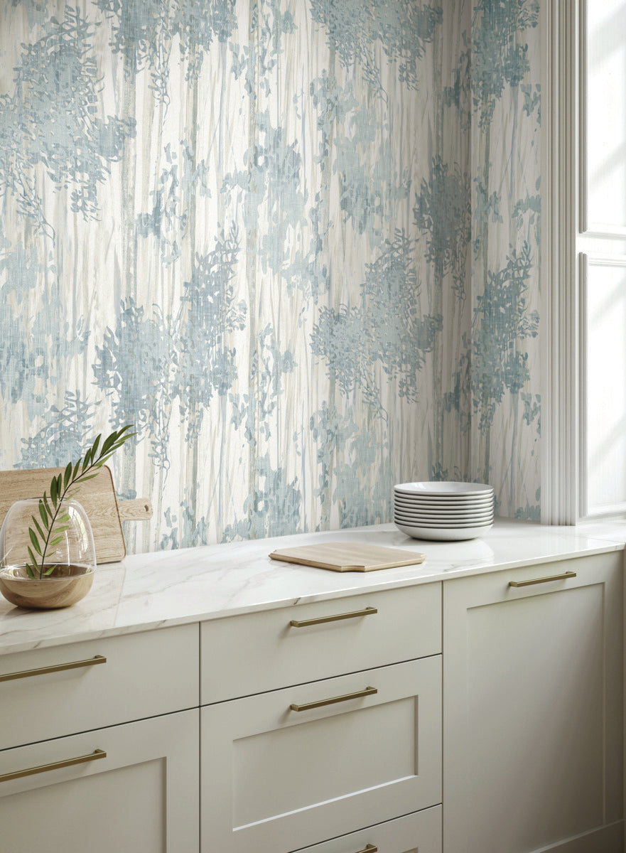 A bright kitchen with white cabinets, gold handles, marble countertop, stacked plates, a wooden cutting board, and a vase with a green branch. The wall features Aspen Grove Blue Wallpaper by York Wallcoverings in an abstract leafy pattern.