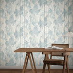 A wooden desk and a woven-back chair sit before York Wallcoverings’ Aspen Grove Blue Wallpaper (60 SqFt). An open book and modern white lamp rest on the desk, enhancing the wooden floor’s warmth.