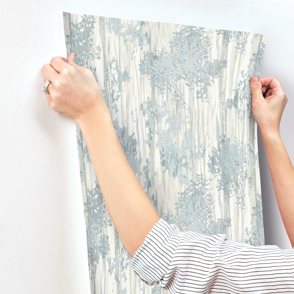 A person with rolled-up sleeves aligns Aspen Grove Blue Wallpaper by York Wallcoverings, featuring blue and white botanical patterns, against a white wall in preparation for application.