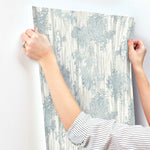 A person with rolled-up sleeves aligns Aspen Grove Blue Wallpaper by York Wallcoverings, featuring blue and white botanical patterns, against a white wall in preparation for application.