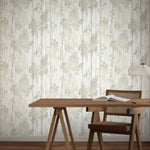 A wooden desk with an open book and a woven chair sits against a wall covered in York Wallcoverings' Aspen Grove Grey Wallpaper (60 SqFt), featuring a subtle abstract pattern. A modern table lamp with a pleated shade is on the right side of the desk.