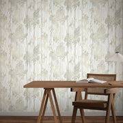 A wooden desk with an open book and a woven chair sits against a wall covered in York Wallcoverings' Aspen Grove Grey Wallpaper (60 SqFt), featuring a subtle abstract pattern. A modern table lamp with a pleated shade is on the right side of the desk.