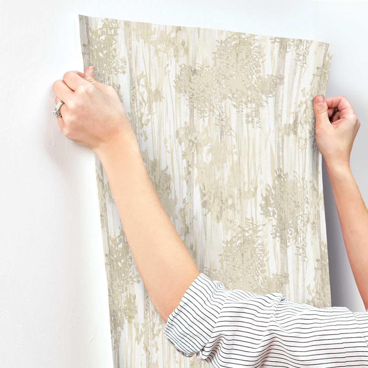 With a ring on their left hand, a person smooths York Wallcoverings Aspen Grove Sand Wallpaper (60 SqFt)—a subtle, leafy beige design—onto a white wall, carefully pressing the pattern into place.