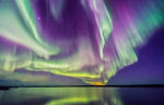 A vibrant display of the northern lights, featuring waves of green and purple auroras across a star-filled night sky, over a dark, silhouetted landscape with a tranquil blue lake from Decor2Go Wallpaper Mural.
