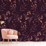 A cozy living room corner features a light pink armchair with cushions against a dark floral wallpaper backdrop. The Decor2Go Wallpaper Mural's Autumn Floral design showcases a pattern of delicate, stylized leaves and flowers in shades of purple and peach. A gold floor lamp stands beside the chair.