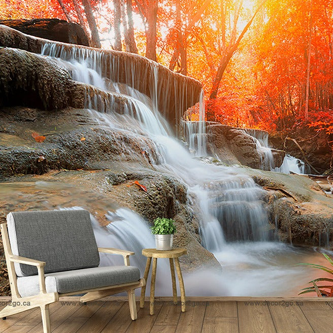 A serene waterfall cascading over rocky tiers in a vibrant autumn forest, juxtaposed with a cozy armchair and side table on a wooden floor, blending outdoor and indoor elements in the Autumn Waterfall Wallpaper Mural by Decor2Go Wallpaper Mural.