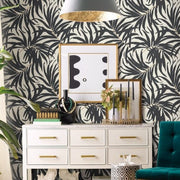 The stylish interior showcases a white dresser with gold handles, adorned with books, vases, and flowers. Above it hangs a framed abstract art piece set against the Black Bali Leaves Wallpaper by York Wallcoverings. A teal chair and a pendant light enhance the nature-inspired design.
