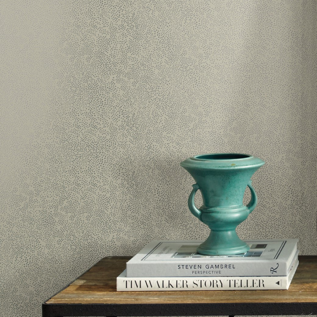 A small turquoise vase with handles sits on stacked books atop a wooden surface, set against York Wallcoverings' Beige Champagne Dots Wallpaper (60 SqFt) for a playful yet elegant touch.