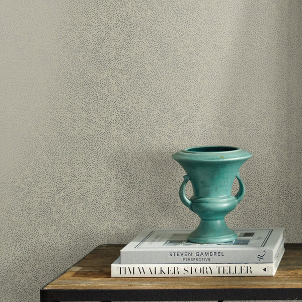A small turquoise vase with handles sits on stacked books atop a wooden surface, set against York Wallcoverings' Beige Champagne Dots Wallpaper (60 SqFt) for a playful yet elegant touch.