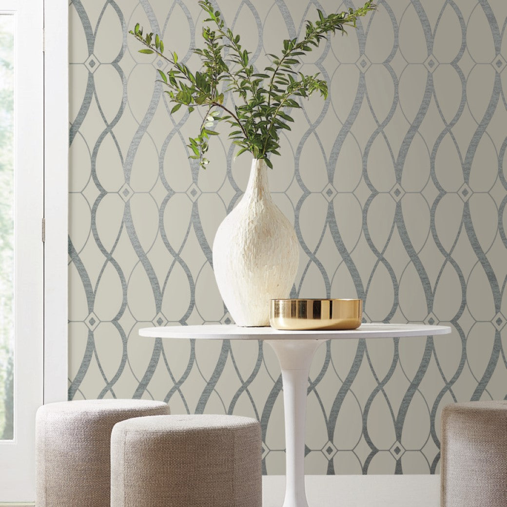 A modern interior showcases a round white table with a textured vase containing leafy branches. A small metallic gold bowl complements this, while two beige upholstered stools encircle the table. The backdrop features York Wallcoverings' Beige & Silver Graceful Geo Wallpaper with a geometric pattern.