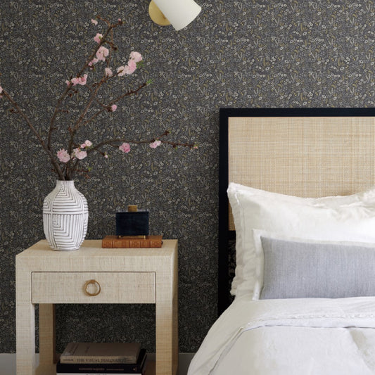 A beige bed with white and gray pillows is next to a wooden nightstand, which holds a textured white vase with flowering branches, books, and a black container. York's Black/Beige Tapestry Wallpaper adorns the wall, while a small light fixture glows above.