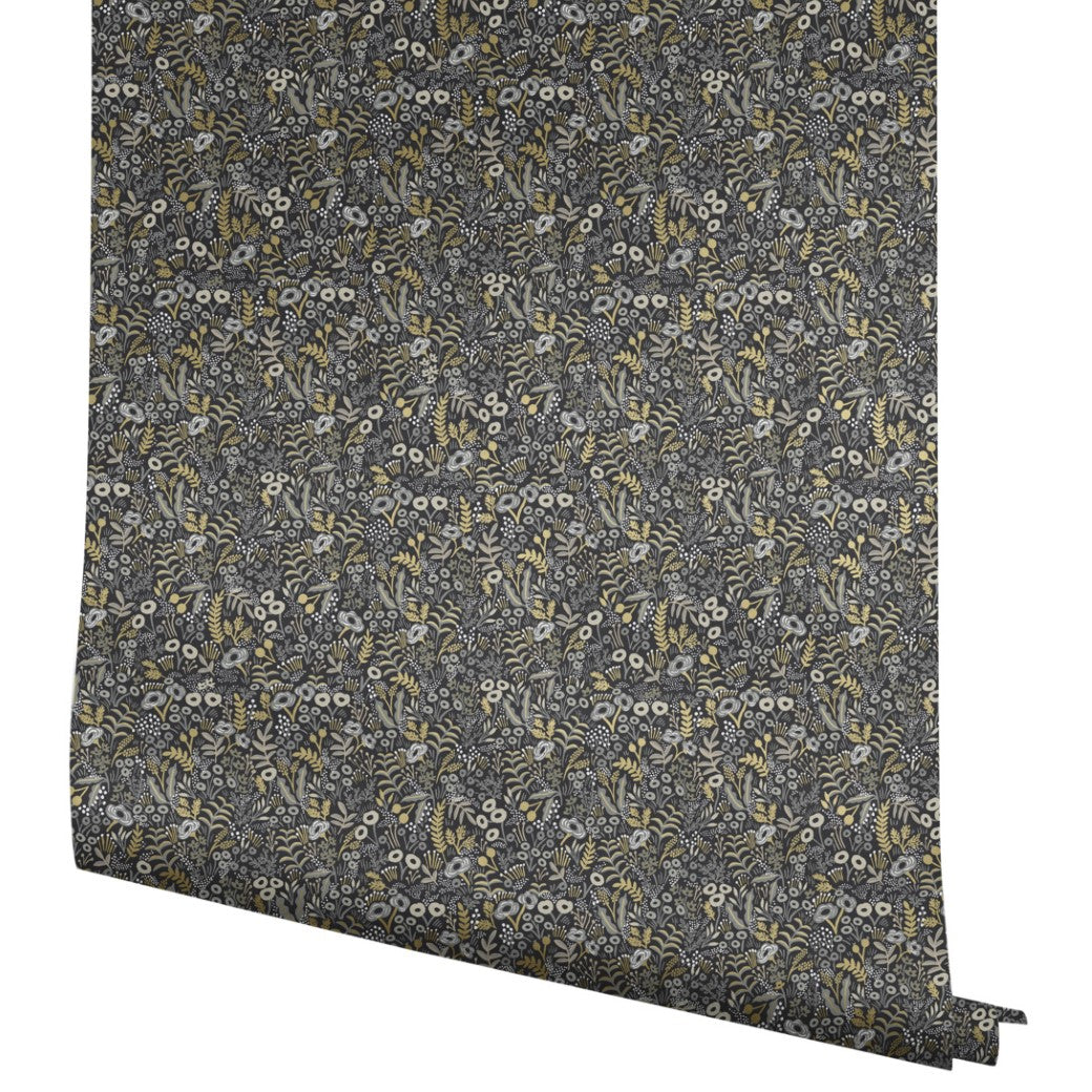 A roll of York Wallcoverings Black/Beige Tapestry Wallpaper (60 SqFt) showcases a dense botanical pattern with small white flowers, yellow accents, and green leaves on a dark background, partially unrolled to reveal its intricate design.