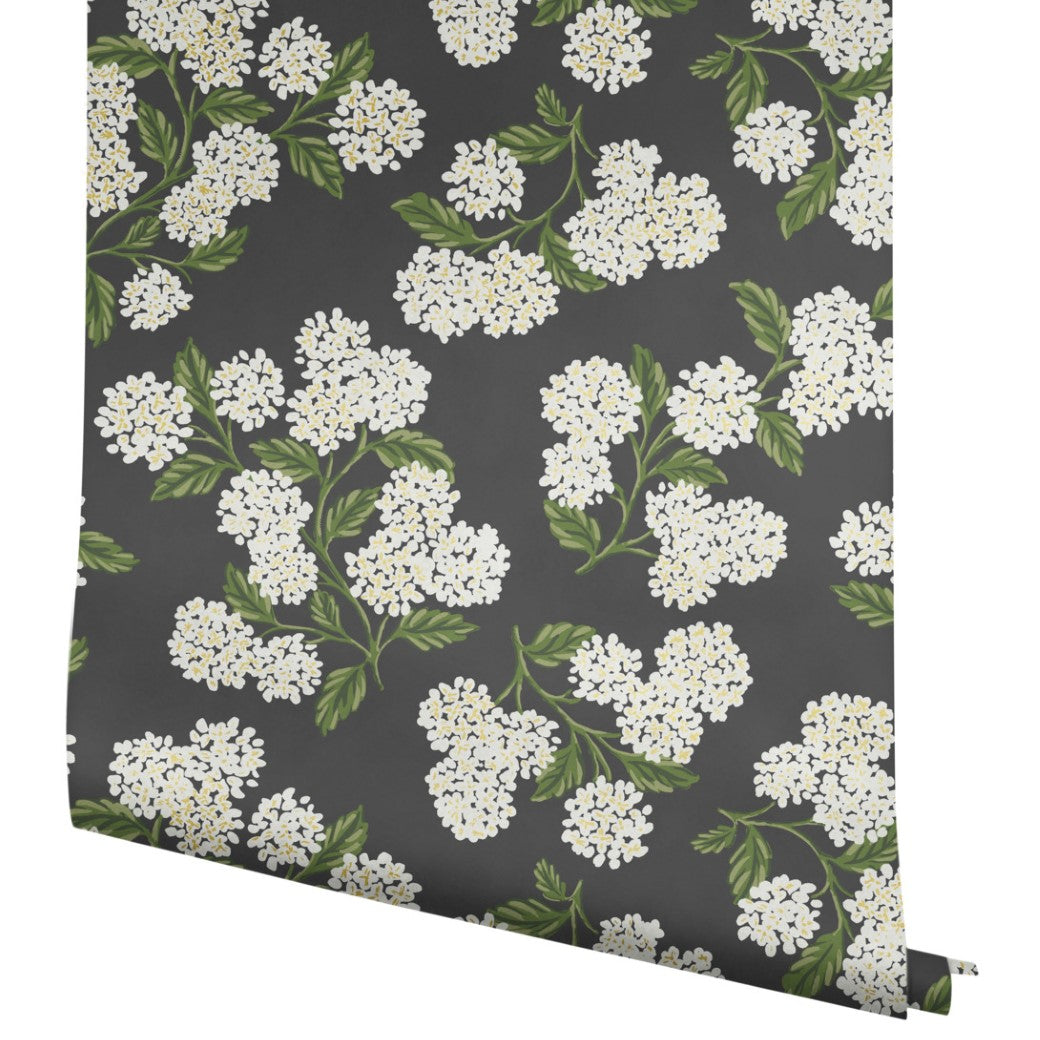 A roll of York Wallcoverings Black/White Hydrangea Wallpaper (60 SqFt) features a dark gray background with white and yellow floral clusters and green leaves in a repeating pattern.