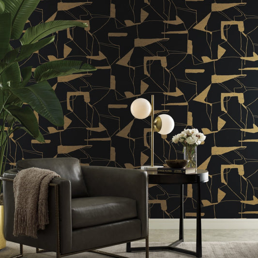 A modern living space with York Wallcoverings' Black & Gold Abstract Geo Wallpaper, a dark gray armchair with a throw blanket, and a black side table featuring metallic elements like a lamp, flowers, books, plus a large green plant on the left.