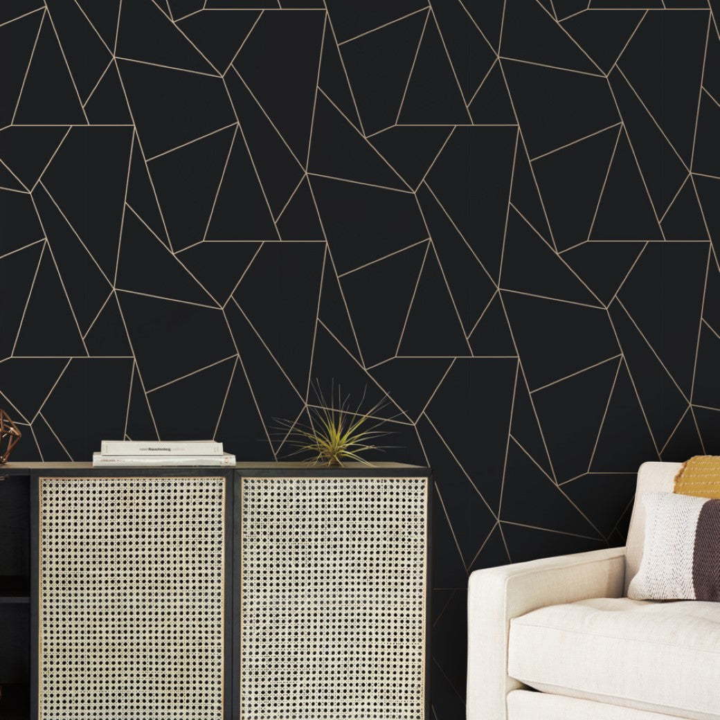 A modern living room showcases a beige couch with a striped cushion. A woven-design cabinet stands against a black wall adorned with York Wallcoverings' Black & Gold Nazca Wallpaper, featuring geometric patterns. A small plant on the cabinet adds greenery to the space.