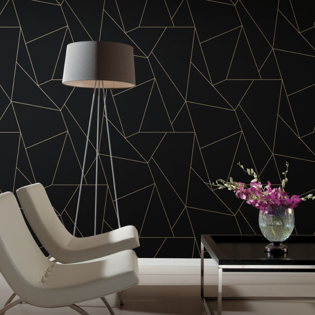 A modern living room with two white chairs and a tall floor lamp features the York Wallcoverings Black & Gold Nazca Wallpaper, showcasing geometric metallic gold lines on a black background. A black table with a vase of purple orchids adds color to the minimalist decor.